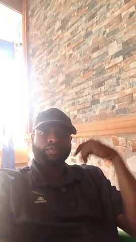A man wearing a black cap and black shirt is sitting down indoors. He has a beard. His left arm is resting on the back of the chair. The background is a brick wall with a window on the upper left. Sunlight is streaming through the window. Music is playing in the background.