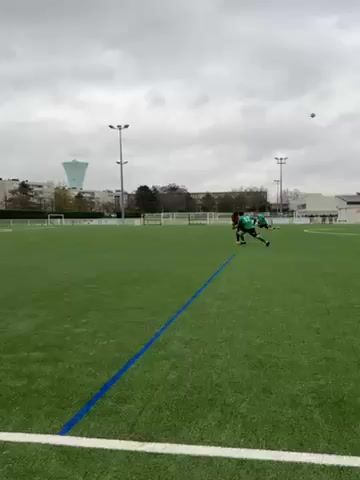 The video depicts a soccer game on a green artificial turf field under a cloudy sky. Several soccer players wearing green and red jerseys are visible. The camera follows a player in red dribbling the ball towards the goal, challenged by a player in green. Buildings, trees, and light posts are visible in the background. White lines mark the field, and a blue line is in the foreground.