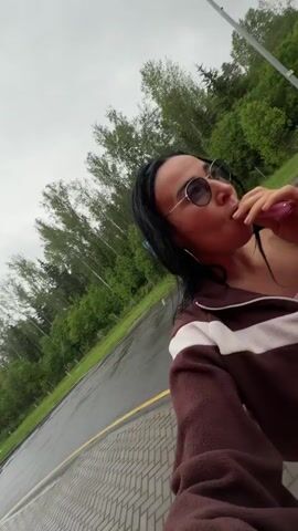 A woman with long black hair and sunglasses is standing on a wet paved road, exhaling a cloud of smoke. She wears a brown and white zip-up fleece jacket over a pink top. Behind her, there are green trees and a parking sign. The sky is overcast.