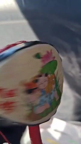 The media file displays a close-up shot of a toy drum, likely for a child, being played. The drum has a rounded shape with a handle. The surfaces of the drum are decorated with colorful cartoon characters, appearing to be from a fairy tale or animated movie. The colors are bright and vibrant. The drum is being spun, indicated by the blur in some frames, causing the attached beads or small mallets to strike the sides of the drum, producing a rhythmic sound.

The background is out of focus, but appears to be an outdoor setting with a grey, textured surface that could be a tent or tarp, possibly illuminated by sunlight casting shadows. A white object, possibly a box or other toy, is visible in the lower right corner. The lighting suggests it's daytime, possibly sunny. The overall mood seems playful and cheerful, associated with children's activities. No text is clearly visible on the drum or in the background. The location context is Glāžšķūnis, Latvia.