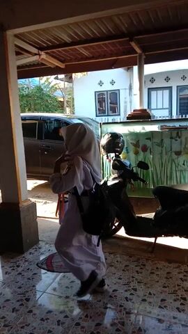 A person wearing a white uniform and hijab is standing in a covered outdoor area with a tiled floor. The person is holding a water bottle and an orange object. A motorcycle is parked behind them, and a car is visible in the background. The area appears to be a driveway or patio attached to a building. The video is shot from a low angle and is rotated sideways.