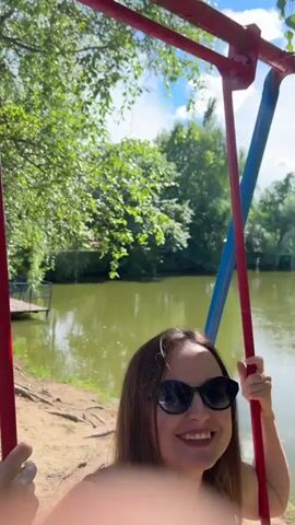 A woman with shoulder-length brown hair and sunglasses sits on a swing by a lake. She is wearing a black tank top and green pants. She is holding onto the swing's chains, which are red and blue. The lake is surrounded by trees, and there is a small wooden dock in the water. The woman is speaking, but her words are not transcribed. She appears to be talking to someone off-camera.
