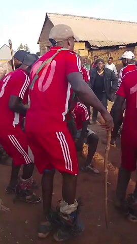 A group of people wearing red jerseys with white numbers and stripes are dancing outdoors in a dirt area. A building with a weathered roof is in the background. One man in the center is squatting, while the others dance around him. One of the dancers is wearing some kind of noisy instrument around their ankles. The group is surrounded by other people, some wearing hats and traditional clothing. The dancers hold what appear to be short wooden poles.