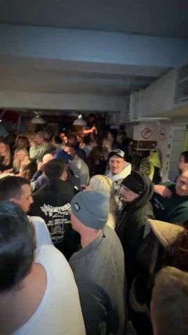 The video shows a very crowded, dimly lit indoor space, seemingly a bar or a club, with a large number of people standing closely together. Many of the people are looking in different directions, and it appears that they are listening to music, as singing can be heard. Some individuals are wearing hats or hoodies. The walls are a light color, and a few signs or decorations are visible in the background. The scene is dynamic and active, with movement and interaction among the people present.