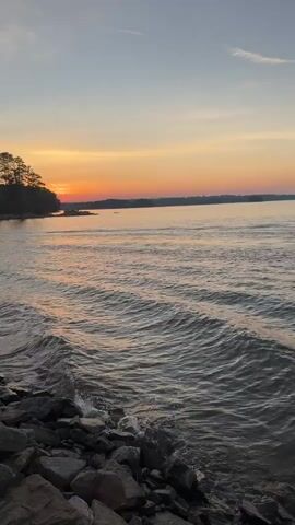This is a vertical video of a sunset over a body of water. The sun is low on the horizon, casting a warm orange and yellow glow across the sky. There are trees on the left side of the frame, silhouetted against the sunset. The water is rippled, with small waves lapping at a rocky shore in the foreground. The sky is a gradient of colors, from the bright sunset hues to a soft blue at the top of the frame. There are no people or objects visible in the water or on the shore. The overall mood of the video is peaceful and serene.