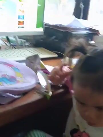 A young girl with her hair in two pigtails, wearing a pink shirt, is sitting at a desk in front of a computer. The computer screen displays a cartoon with a bear and other characters. The girl is looking at the camera and appears to be speaking or making a noise. There are papers and other items on the desk. The scene is indoors, with a window in the background.