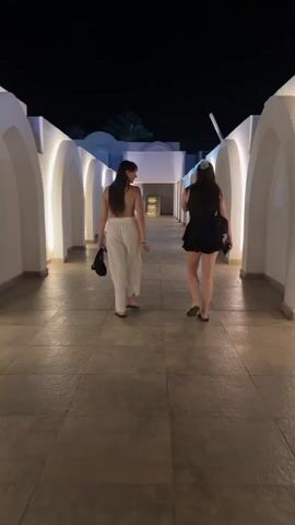 Two women are seen from behind, walking away from the camera down a well-lit outdoor corridor at night. The walkway features repeating white arched structures on both sides, which are illuminated from within by warm-toned lights. The ground is paved with light-colored tiles.

The woman on the left has long, dark hair and is wearing a light-colored, possibly white, bra top or crop top with matching loose-fitting pants. She carries a small black handbag in her right hand and is wearing dark flip-flops.

The woman on the right also has long, dark hair and is dressed in a dark, short dress or romper. She carries a larger dark bag on her left shoulder and is wearing dark flip-flops.

In the distance, beyond the corridor, the dark night sky is visible, along with what appears to be a palm tree, suggesting a warm climate or resort environment. The overall ambiance is tranquil and inviting due to the soft lighting. No text is visible.