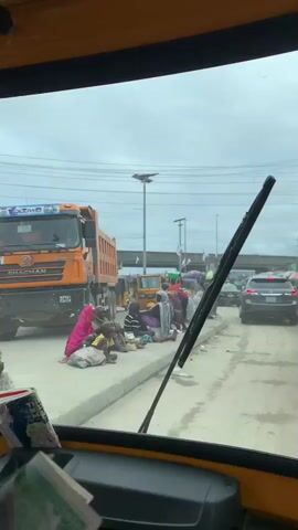 The video, shot from the perspective of the passenger in a yellow motor tricycle, shows a group of people sitting on the curb of a busy street. The group consists of both adults and children. They appear to be sorting through items laid out on the ground in front of them, possibly collecting or organizing items. An orange dump truck is parked nearby on the left side of the street. Various other vehicles, including motorcycles and cars, pass by on the street. The sky is overcast. The passenger takes a mirror selfie in the right-side mirror of the tricycle. The scene suggests an urban area, possibly in a developing country.