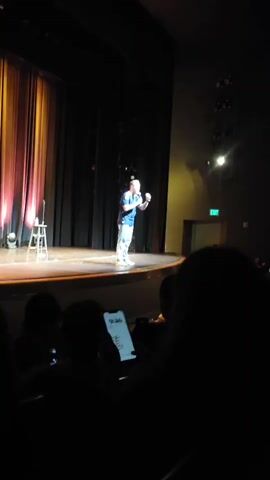 A man is on stage in a theater, standing to the right of a stool with a microphone. He is wearing a blue t-shirt and white pants. The stage has a curtain behind it with red and yellow lighting. The audience is visible in the foreground, with some people holding up phones. The man is gesturing with his right hand.