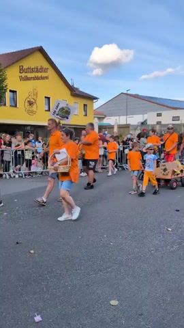 A group of people in orange shirts are walking down a street, some are carrying baskets and others are pulling small wagons. There are spectators on the side of the street. A yellow building with "Buttstädter Vollkornbäckerei" written on it is visible on the left side of the frame. The sky is blue with some clouds. The scene appears to be a parade or a community event.