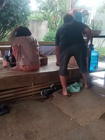 In the video, a man in a black shirt and gray shorts stands on a wooden platform next to a table with a metal container, seemingly preparing food or drinks. He faces away from the camera, looking towards an area with trees and greenery. A woman, dressed in a tan shirt and floral pants, is squatting on the platform, also facing the greenery. Another man, wearing a gray shirt, sits crouched down next to her, interacting with something on the platform. Shoes are visible to the right of the platform, on a concrete surface. The environment suggests an outdoor or semi-outdoor setting, possibly a porch or sheltered area, with a lush background.