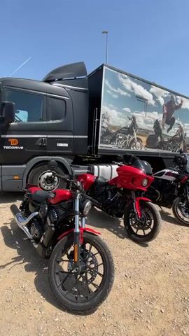The video shows a row of Harley-Davidson motorcycles parked on a gravel surface in front of a large black truck with a trailer. The trailer is decorated with an advertisement for the "Harley-Davidson Experience Tour" featuring images of motorcycles and a rider in the air. The motorcycles are of different models and colors, including red and black. The sky is blue and clear. There is a street lamp visible in the background. The overall setting appears to be an outdoor event or promotional display.