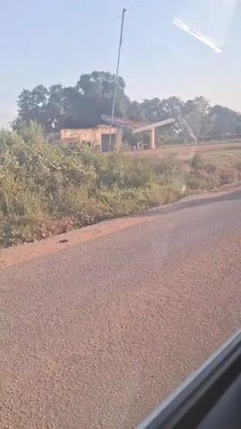 The video shows a road in a rural area on a sunny day. A motorcycle loaded with several large white sacks drives past. In the background is an old, partially ruined building surrounded by overgrown vegetation. A tall pole stands nearby. The video is shot from inside a moving vehicle, showing the passing landscape.
