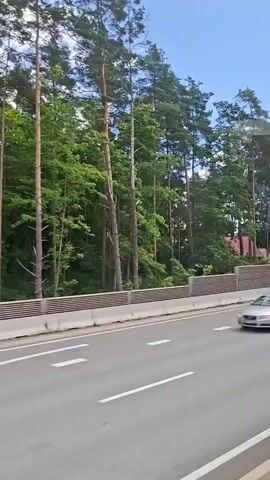 A video captures a view from a moving vehicle, likely a bus or car, as it travels along a road. The road is paved and marked with white lines, indicating multiple lanes. A sound barrier made of horizontal slats is visible alongside the road. Beyond the barrier, a dense forest of tall pine trees grows, with glimpses of red-roofed buildings nestled within the trees. The sky is blue with scattered white clouds. The vehicle continues to move, revealing a wider view of the road as it curves. In the distance, a modern building with "Sanitex" written on its facade is visible, surrounded by green grass and trees. Streetlights and road signs are also present. The scene is peaceful and appears to be a sunny day in a semi-rural or suburban area.