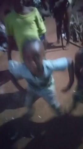 The video shows a group of children in what appears to be a dirt clearing at night. One child is in the foreground wearing a light-colored shirt with a graphic and light-colored pants. This child is dancing with energy, moving their arms and body in a rhythmic way. Other children are standing around, some watching and some partially obscured. The light is dim, and there are shadows around the children. Some bits of debris or small objects are visible on the ground. In the background, there are what seem to be wood pieces scattered. The focus is mainly on the child dancing.