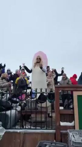 The video captures a festive scene on what appears to be a wooden deck or balcony, possibly at a ski resort, given the winter attire of the crowd. The sky is overcast. A group of people are gathered, many holding drinks, seemingly celebrating. The focal point is a person dressed in an inflatable costume resembling a marshmallow or similar confection, complete with a pink top. This person stands on a metal table or platform, holding a wine glass and dancing enthusiastically, while the crowd cheers and raises their glasses in unison. There are metal railings and snow visible on the ground, adding to the winter atmosphere. Some ski gear is also present.