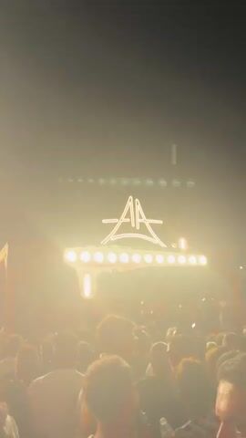 The video shows a crowded outdoor nightclub at night. A neon sign with the letters "AA" in a stylized font illuminates the scene from above a wooden structure. Below the sign, a DJ booth with red lighting is visible, with people dancing in front of it. The crowd is diverse, with individuals of various ages and genders, all appearing to be enjoying the music and atmosphere. The lighting is dynamic, with bright spotlights and colorful beams illuminating the crowd. The overall impression is one of a lively and energetic party.