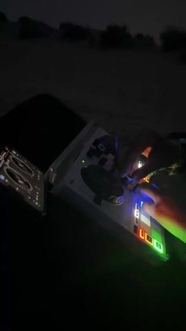 In the low-lit video, a person is shown operating a DJ controller with illuminated buttons, accompanied by an open tablet displaying DJ software. The music emanating from the controller is a heavy techno beat. The camera then pans up, revealing a dark beach scene with silhouettes of beach umbrellas against the night sky. The environment is dimly lit, emphasizing the contrast between the illuminated DJ equipment and the darkened landscape.