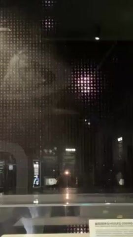 Here's a detailed description of the video:

The video shows a display behind glass. The display is a grid of letters, specifically the DNA bases A, T, C, and G, arranged in a dense pattern.  Reflections are visible on the glass, showing what appears to be an indoor setting, possibly a museum or exhibition. Lights and other displays are visible in the reflections.  A sign with text in a non-English language (likely Chinese) sits below the glass display.  The lighting is dim, with purple hues emanating from the displays behind the glass.