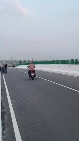 This is a vertical video clip of an elevated highway in Rupganj, Bangladesh. The sky is overcast, suggesting either early morning or late afternoon. The highway itself is paved and has clear white lane markings. White concrete barriers line the right side of the road, with a green fence behind them. Streetlights are visible on poles along the left side of the road.

The primary subjects are people on motorcycles and walking. In the foreground, a person in a pink shirt rides a motorcycle away from the camera. Further down the road, another motorcycle with a person wearing a dark hijab is parked on the left side, and two people are walking past it. Several other figures are visible further in the distance, also on foot.

In the background, there are trees and some buildings, including a distinctive red and white tower or structure. The overall scene conveys a sense of a new or developing infrastructure project, with the open highway and the people utilizing it. There is no visible text in the video.