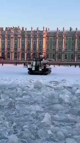 The video shows a black airboat maneuvering over a snow-covered area covered in ice chunks. In the background, there is a large ornate building with many windows. People are walking in the snow near the building. The airboat kicks up a cloud of snow as it moves. The sky is blue.