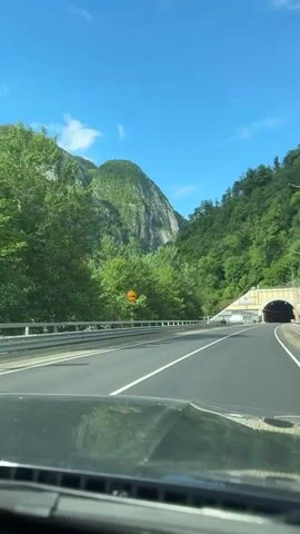 From the perspective of a moving vehicle, the camera captures a scenic drive towards a tunnel on a clear, sunny day. The road, marked with solid white lines, leads directly to the tunnel entrance, which is painted with yellow and beige stripes. A man stands on the side of the road wearing a white shirt and holding a high-visibility yellow vest. The surrounding landscape features lush green mountains covered in dense trees. The sky is bright blue, with a few scattered clouds. The vehicle's dashboard is visible in the foreground.