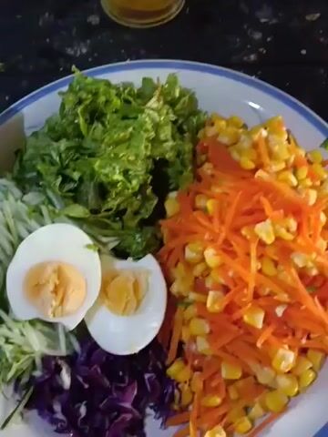 The video shows a close-up of a salad on a plate. The salad contains lettuce, shredded carrots, corn, shredded cucumber, red cabbage, and two halves of a hard-boiled egg. A bottle of Maestro brand salad dressing, labeled "Wijen Sangrai," is placed next to the salad. In the background, there are bags of colorful snacks. The lighting suggests an indoor setting. The camera is relatively still, with some slight movement. The video appears to be a product shot or a food preparation showcase.