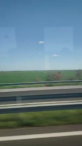 The video shows a view from a moving vehicle, likely a car or bus, traveling along a highway. The landscape is predominantly flat farmland under a clear blue sky with a few scattered clouds. A metal guardrail runs alongside the highway, and there is green vegetation on the side of the road. The movement indicates the vehicle is in motion.