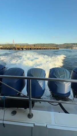 A POV shot from a boat shows three blue outboard motors covered with dark blue protective covers. The boat is moving forward through the water, creating a large white wake. In the distance, a wooden pier stretches out into the water, with a few buildings and trees visible on the shore behind it. The sky is clear and blue.