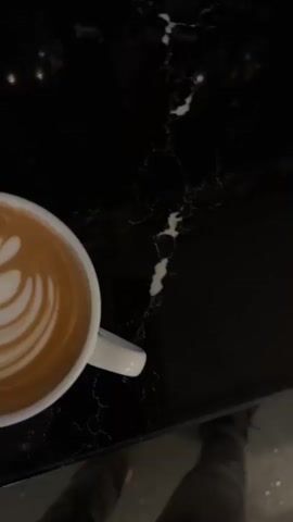 This is a close-up, top-down video of a white mug of latte art on a black marble surface. The latte art features a symmetrical pattern of white foam resembling a leaf or a tulip with small hearts at the top. The camera slowly zooms in and out, and the lighting shifts slightly, causing subtle changes in the sheen of the marble and the coffee. No people are visible in the frame. The overall impression is one of stillness and appreciation for the aesthetic of the coffee.