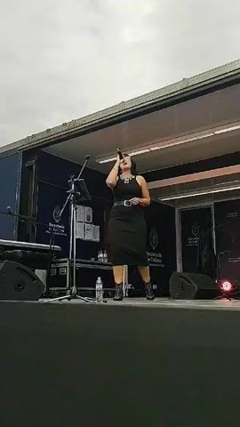 A woman is singing on a stage. She is wearing a black dress, a belt, and a statement necklace. She has short black hair and is wearing glasses. She is holding a microphone and singing. There is a microphone stand and a water bottle next to her. Behind her is a black backdrop with the logo for the Secretaría de Cultura, Presidencia de la Nación. There are speakers and other equipment on the stage. The sky is overcast.
