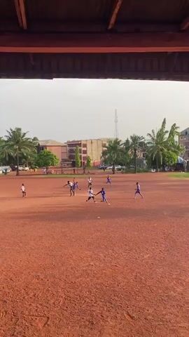 A video shows a soccer match being played on a red dirt field, viewed from under a covered structure. Players in white and blue jerseys run across the field, chasing a soccer ball. Several palm trees line the edge of the field in the background, interspersed with buildings and a telecommunications tower. At the end of the video, the back of a person's head is visible, suggesting they are also watching the game.