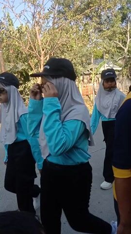 A group of people, some wearing matching uniforms with blue and yellow accents, stand on a paved surface outdoors. One person in the foreground, wearing a yellow shirt with blue sleeves and a dark headscarf, is partially visible and appears to be looking down. Behind them, another individual in a grey and blue uniform, also wearing a headscarf and a cap, stands with their legs spread apart, facing away from the camera. Further back, another person in a similar uniform is visible. A motorcycle drives past in the background. The environment is characterized by trees and vegetation.
