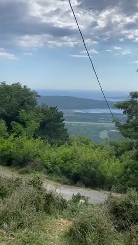 A scenic view of a bay and coastline is seen from a hilltop overlooking a lush green landscape. A thin black cable runs diagonally across the frame from the top left to the bottom right. In the foreground, a dirt path winds its way down a slope covered in dense green bushes and dry grasses. Beyond the path, a thick forest of green trees covers the hillside. In the mid-ground, the landscape opens up to reveal a body of water, likely a bay, with a distant coastline stretching across the horizon. A thin strip of land, possibly an airport runway, is visible near the water's edge. The sky above is a bright blue, dotted with fluffy white clouds, suggesting fair weather. The overall scene evokes a sense of tranquility and natural beauty.