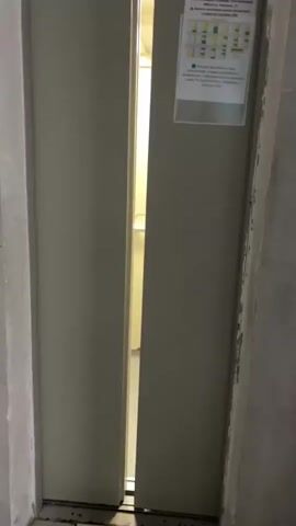 The video begins with a view of an elevator with its doors slightly ajar. The interior of the elevator is visible, showing a mirror on the back wall. As the doors open further, the camera moves inside the elevator. The elevator is small and utilitarian, with cream-colored walls and a gray floor. A security camera is mounted on the ceiling, and fluorescent lights illuminate the space. A handrail runs across the mirror. On the left wall, there are several posters or notices. A person, seen in the mirror, is standing in the elevator, holding a phone and appearing to take a selfie. They are wearing a light-colored t-shirt with a graphic print and dark shorts. Behind the person, also reflected in the mirror, are numbers arranged vertically on a teal-colored wall. The elevator appears to be in a building, possibly an office or residential complex. The video shows the elevator doors opening, the interior, and a person taking a photo of themselves.