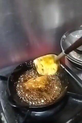 A cooking utensil with a wooden handle is holding a piece of food that is submerged in a bubbling liquid within a black frying pan. The pan is on a stovetop with a metallic backsplash behind it.