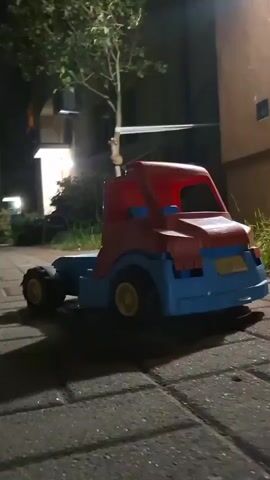A toy semi-truck is shown in close-up, with the camera moving slightly. The truck is red and blue with yellow wheels. It is on a paved walkway at night. There is a tree to the left and a building with lights and plants to the right. The truck appears to be stationary.