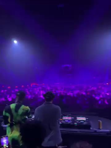 The video captures a dynamic DJ set taking place at a large music event in Indio, United States. The scene is enveloped in a dark, expansive venue, brightly illuminated by intense purple and blue stage lights and sweeping laser beams, creating an energetic and immersive atmosphere.

In the foreground, two DJs are visible from behind, actively performing at their mixing consoles. The DJ on the left wears a distinctive bright green and black patterned shirt, while the DJ on the right is dressed in a light grey or white shirt. Both are wearing headphones, focused on their equipment as they control the music. The DJ setup, featuring multiple turntables and mixers, is clearly visible before them.

Beyond the stage, a vast crowd fills the space, stretching into the distance. The attendees are largely obscured by the vibrant lighting and haze, but their presence is evident, with numerous small lights (likely from mobile phones or light sticks) twinkling throughout the audience, suggesting an engaged and enthusiastic crowd. The overall ambiance is one of high energy and excitement, characteristic of a major music festival or concert at night. No specific text is visible within the frame.