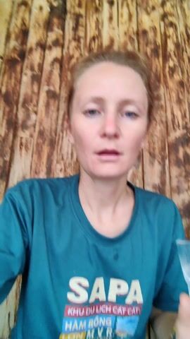 A fair-skinned woman with blonde hair in a bun, wearing a teal t-shirt with "SAPA" printed on it, raises a clear glass of water and takes a sip. The background is a rustic wooden wall. She is wearing a beaded bracelet on her right wrist. The glass has a decorative footed base.