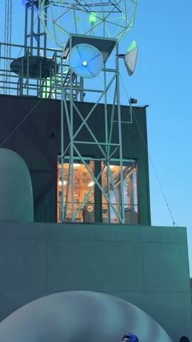 The video shows a building facade with various antenna-like structures atop it, illuminated with changing lights. The structures are built from thin metal framework and circular satellite-dish shapes.  The main building element is grayish-green, with a tower-like structure rising from it.  A rectangular window, illuminated with warm light from the interior, is visible on the building.  A person is seen moving inside this window.  The sky is a clear blue, hinting at either late afternoon or early evening. Clothes seem to hang on one of the metal structures.  The lighting on the antenna-like structures changes from green to blue. Music plays in the background.