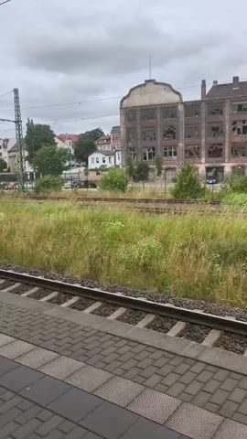 The video captures a view from a train platform, looking out over railroad tracks towards a weathered, multi-story building with many broken windows. The building appears to be industrial or commercial and is surrounded by overgrown vegetation, including tall grass and trees. The sky is overcast with clouds. The train platform itself is paved with brick-like tiles.