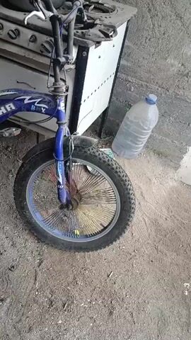 The camera pans around a blue "DECHA" brand BMX-style bicycle resting on a dirt and gravel surface. The bike has fat tires and colorful spokes. Behind the bike, there is a worn, white-and-black stove, a large plastic bottle of water, and some other miscellaneous items including a cooking pot and plastic crates.  The setting appears to be a cluttered outdoor or semi-outdoor area, possibly a shed or garage. There are no people visible in the video.