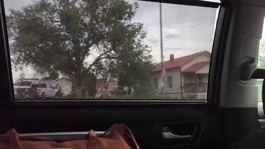 The video shows a view through the window of a moving car. The window has a mesh sunshade. Outside, the scenery consists of houses and trees under a cloudy sky. In the foreground inside the car, a brown bag is visible on the seat. The car appears to be driving down a street in a residential area.