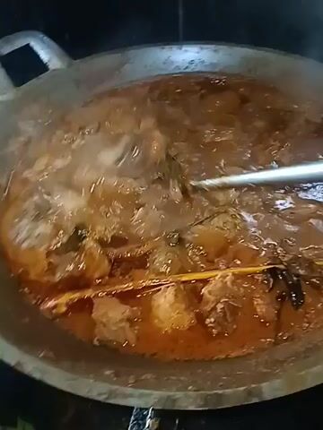 A close-up, overhead shot shows a large, dark, shallow pan filled with a rich, reddish-brown stew. Several pieces of meat, appearing to be chicken or pork, are visible submerged in the liquid, along with what looks like stalks of lemongrass and possibly star anise. Steam rises from the stew, indicating it is hot and actively cooking. A metal spatula or ladle is being used to stir the contents of the pan, creating ripples on the surface of the stew and agitating the steam. The pan itself is dark gray and has a slightly worn appearance. The background is dark and out of focus, suggesting an indoor cooking environment.
