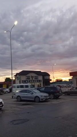 Here's a description of the video:

The video shows a parking lot with several cars parked under a cloudy sky. The light from street lamps illuminates the area. A building with a sign that says "ТАБАК" (Tobacco) in Russian is visible in the background. There are other buildings and structures in the distance. The general impression is that it is dusk or dawn, given the lighting conditions.