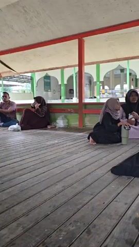 The video shows a group of four women sitting on a wooden floor under a covered shelter with a red roof trim and green pillars. In the background, there is a building with green walls and white arched openings. The women are all wearing hijabs. Two women in the foreground are sitting close together and appear to be talking. One woman is wearing a black hijab and a light purple patterned dress, and the other is wearing a black hijab and a black dress. A metallic water bottle is visible between them. To the left, another woman in a dark red or purple hijab and dress is sitting alone, looking down. To the far right, a fourth woman, mostly obscured by a blue hijab and black garment, is also sitting. A red plastic bag and other items are scattered on the floor. The overall scene appears to be a casual gathering in an outdoor area, possibly a rest stop or waiting area.
