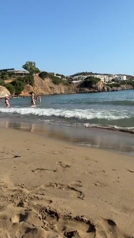 The video shows a beach scene with clear blue skies and calm, wavy ocean water. The sandy beach has footprints visible. In the distance, there are cliffs with buildings on top. Several people are in the shallow water, some appearing to be playing or wading. Closer to the foreground, a young boy wearing a baseball cap and blue shorts is sitting on the wet sand and building a sandcastle. He is focused on his creation, with the sandcastle cone-shaped. The camera perspective is from behind the boy, looking out towards the ocean.