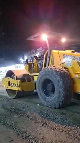 The video shows a yellow Sakai SV526D vibratory roller on a construction site at night. The roller is positioned on a gravelly, uneven surface, likely a road under construction. The large vehicle has prominent headlights illuminating the immediate area, casting bright beams into the darkness. The roller is stationary, and the scene appears to be from a low-angle perspective, emphasizing the size of the machinery.

Visible text on the roller includes "SAKAI" on the front drum and "SV526D" on the engine cover. There's also a green logo on the side of the engine cover, although the details are not clear. In the background, there are blurry lights and structures, suggesting an urban or semi-urban setting, possibly with buildings and streetlights. The overall atmosphere is one of ongoing construction work, carried out at night. The time of day is clearly night due to the artificial lighting and dark sky. The weather is not discernible from the footage. The specific location is identified as Blangpeuria, Indonesia, which provides geographical context to the construction activity. No people are clearly visible in the frame, but there might be an operator inside the roller, obscured by the bright lights and the cab structure. The emotion conveyed is one of industrial activity and the perseverance of construction work, even in the dark.