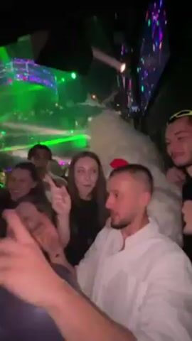 The video shows a group of people dancing in what appears to be a nightclub or party venue. The lighting is dim with green spotlights shining. Several people are visible, including some with balloons. In the center of the group is a person wearing a large, fluffy white costume with red lips. The people appear to be enjoying themselves, dancing and posing for the camera.