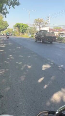 The video shows a two-lane asphalt road on a sunny day, with shadows of trees on the road. On the left side of the road, there is a motorcycle with a rider wearing a green shirt and red helmet. On the right side of the road, there is a truck. The camera appears to be mounted on a moving vehicle, likely a motorcycle given the angle and handlebars visible at the bottom of the frame.